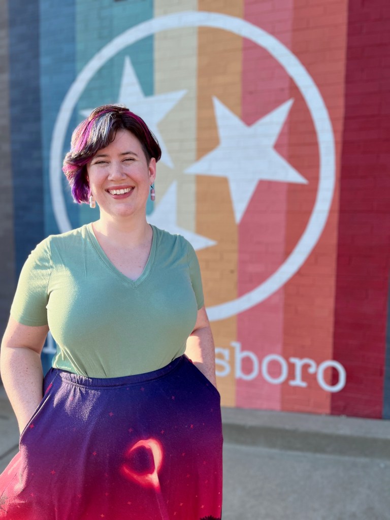 Colleen stands in front of a rainbow Murfreesboro mural and smiles at the camera